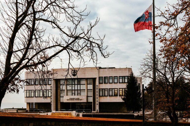 A view of the Slovak Parliament building surrounded by autumn trees with the national flag waving under an overcast sky.