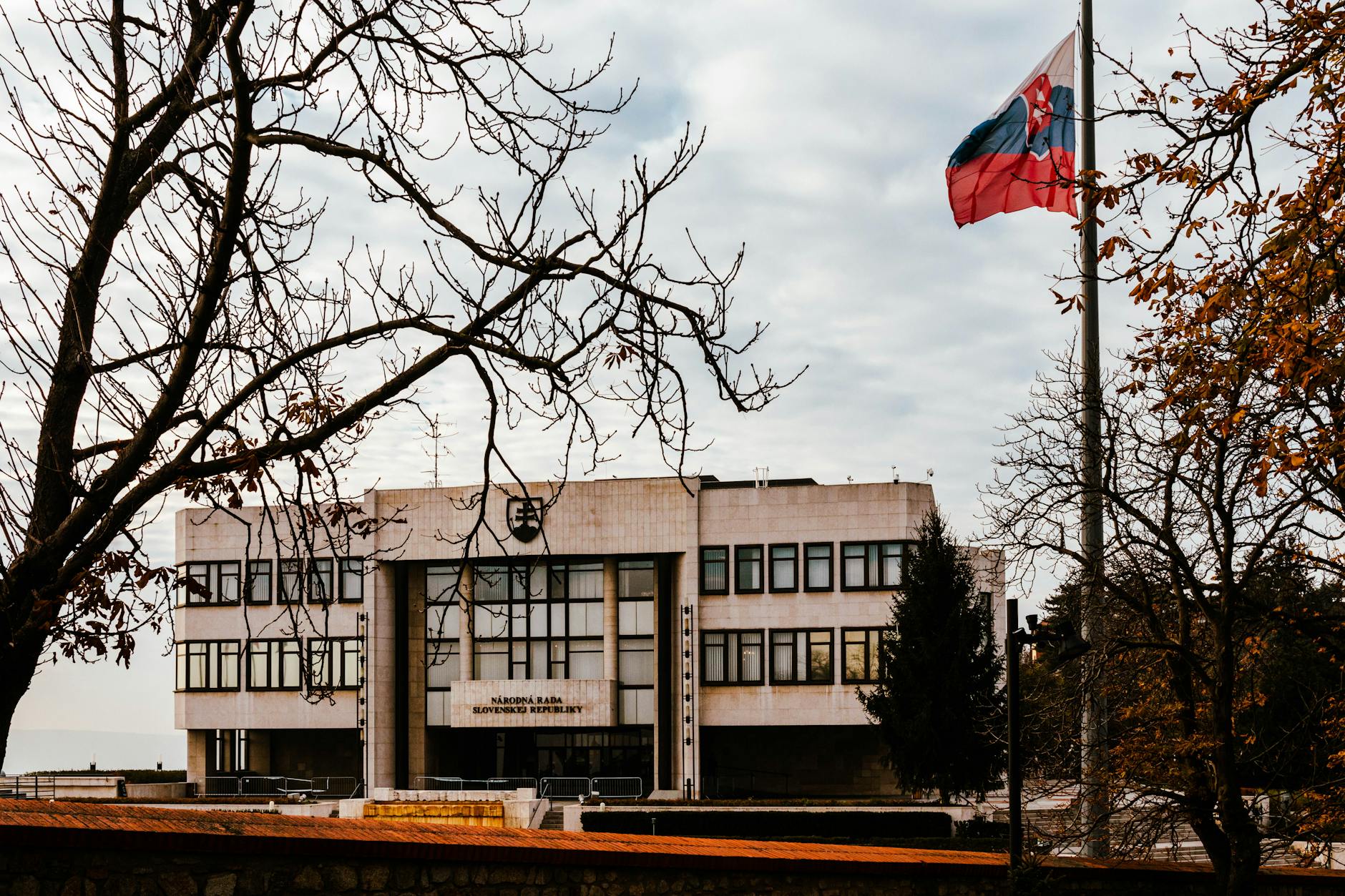A view of the Slovak Parliament building surrounded by autumn trees with the national flag waving under an overcast sky.